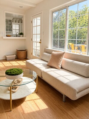 Una sala moderna con un sofá beige, una mesa de centro de cristal con una planta verde, y vistas a sillas amarillas al aire libre a través de amplias ventanas.