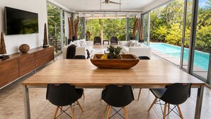 Een moderne eetruimte met een houten tafel, zwarte stoelen en een groot raam met uitzicht op een zwembad en weelderige begroeiing.