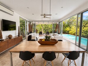Ein moderner Essbereich mit einem Holztisch, schwarzen Stühlen und einem großen Fenster mit Blick auf einen Swimmingpool und üppiges Grün.