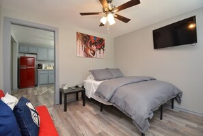 A bedroom with a bed, a nightstand, a ceiling fan, a TV, and a kitchen area visible through an open door.