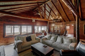 A log cabin interior with a wooden ceiling, large windows, a sofa, a coffee table, and a chandelier. - Rustic Wilson Bay Cabin on Gull Lake | Beach & Dock (Nisswa)