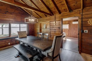 Ein Holzblockhaus im Inneren mit Esstisch, Stühlen, einer Hängelampe und Blick ins Freie.