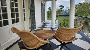 A covered porch with two wicker chairs, a checkered floor, and a view of greenery and a building in the background.