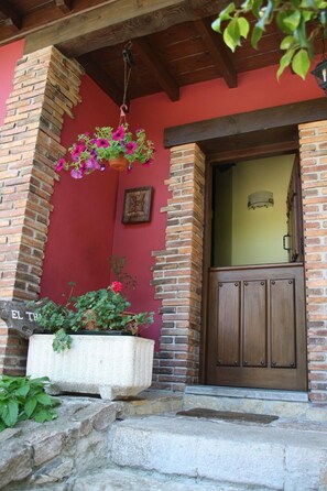 A brick building with a wooden door, a hanging flower basket, and a planter with plants. - Casa Rural El Trechal (Pilona)