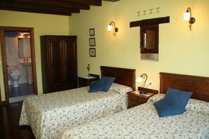 A room with two beds, wooden headboards, a nightstand with a lamp, a small table with a vase, and a bathroom visible through an open door.