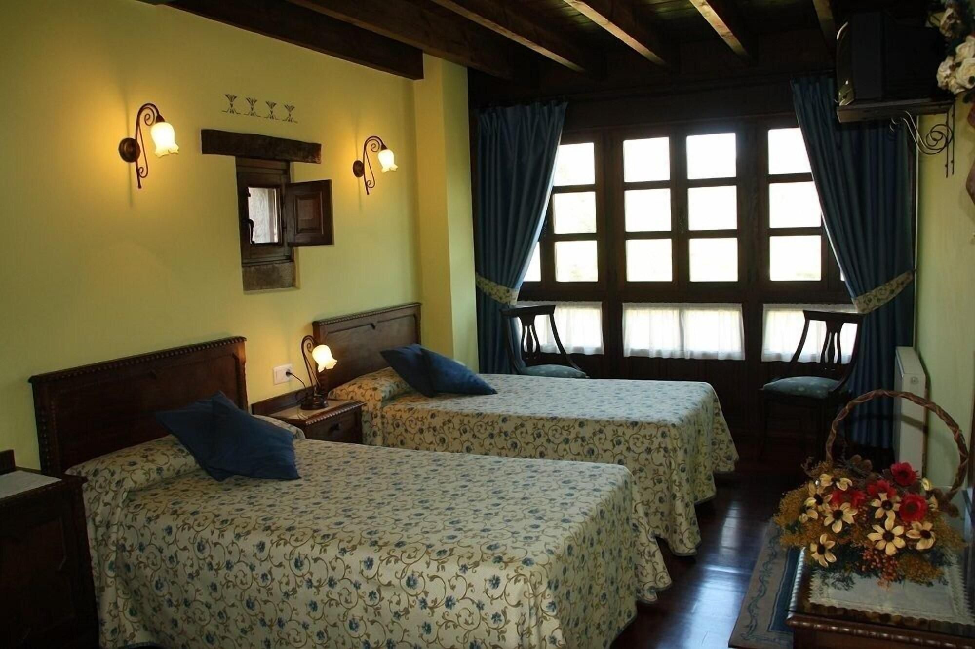 Una habitación con dos camas, techo de madera, ventana con cortinas y una lámpara fijada a la pared.