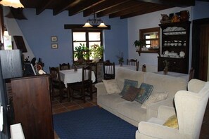 A living room with a sofa, a dining table, chairs, a bookshelf, and a window with plants.