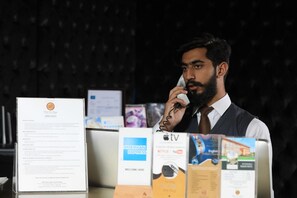 A man at a hotel reception desk with a phone, a policy document, and various informational brochures. - Bella View Lounge (Islamabad)