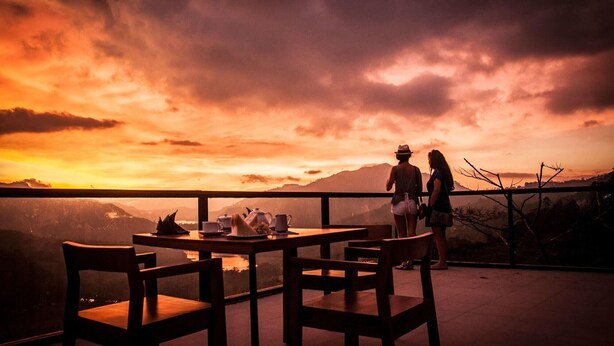 Una terraza con sillas y mesas de madera, con vistas a un paisaje montañoso al atardecer.