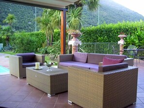 An outdoor patio with wicker furniture, a decorative urn, and a view of a lush garden and mountains. - Hotel Del Sole (Sora)