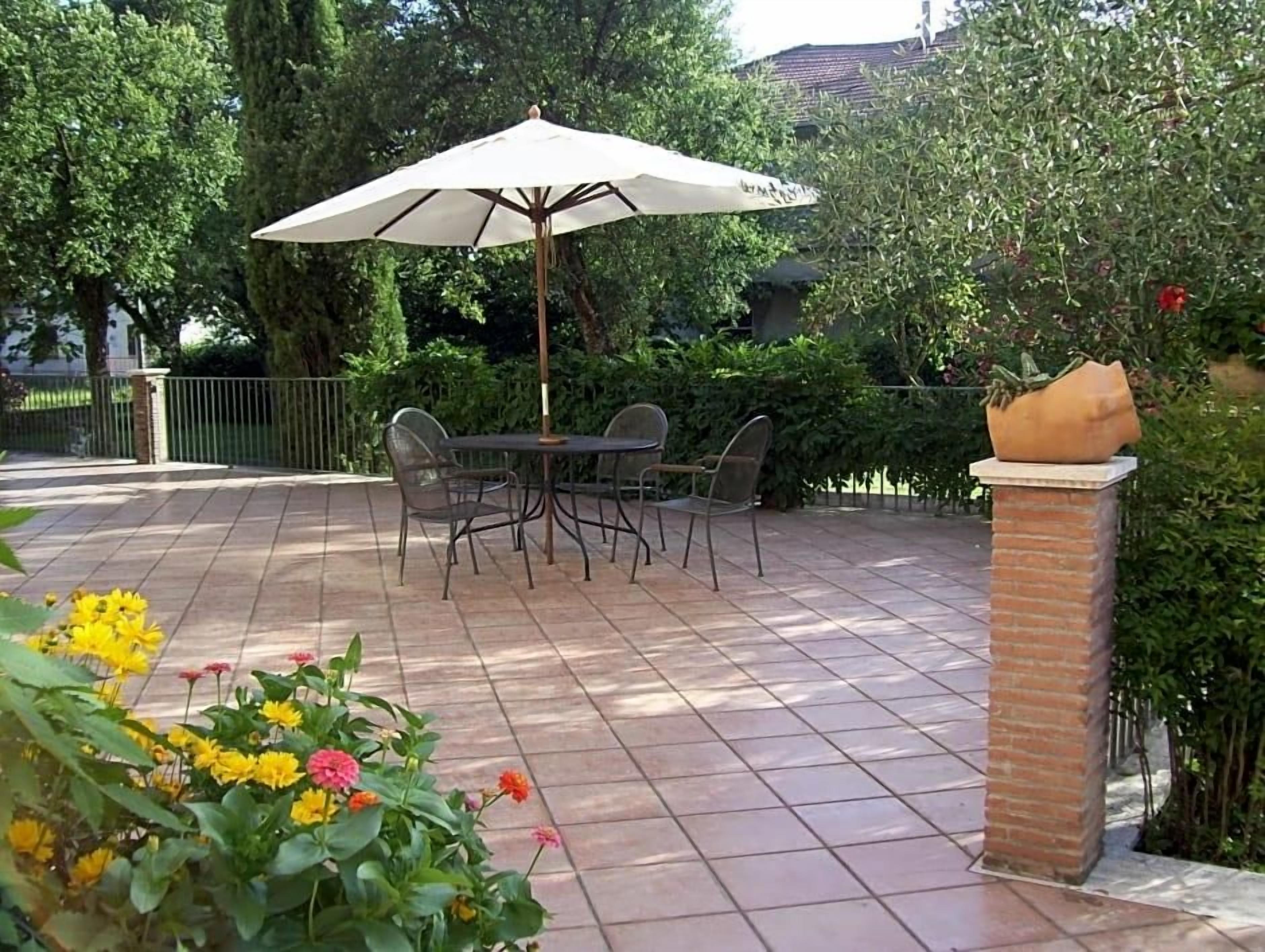 Eine Terrasse im Freien mit Tisch und Stühlen unter einem großen Sonnenschirm, umgeben von viel Grün und einer Ziegelstütze.