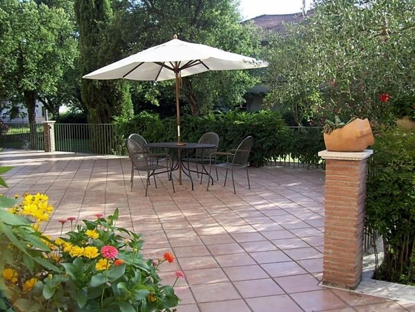 An outdoor patio with a table and chairs under a large umbrella, surrounded by greenery and a brick pillar. - Hotel Del Sole (Sora)