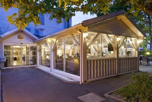A covered outdoor seating area with glass walls, a wooden deck, and a building entrance with sliding glass doors. - Hotel Acadie Les Ulis (Villebon-sur-Yvette)