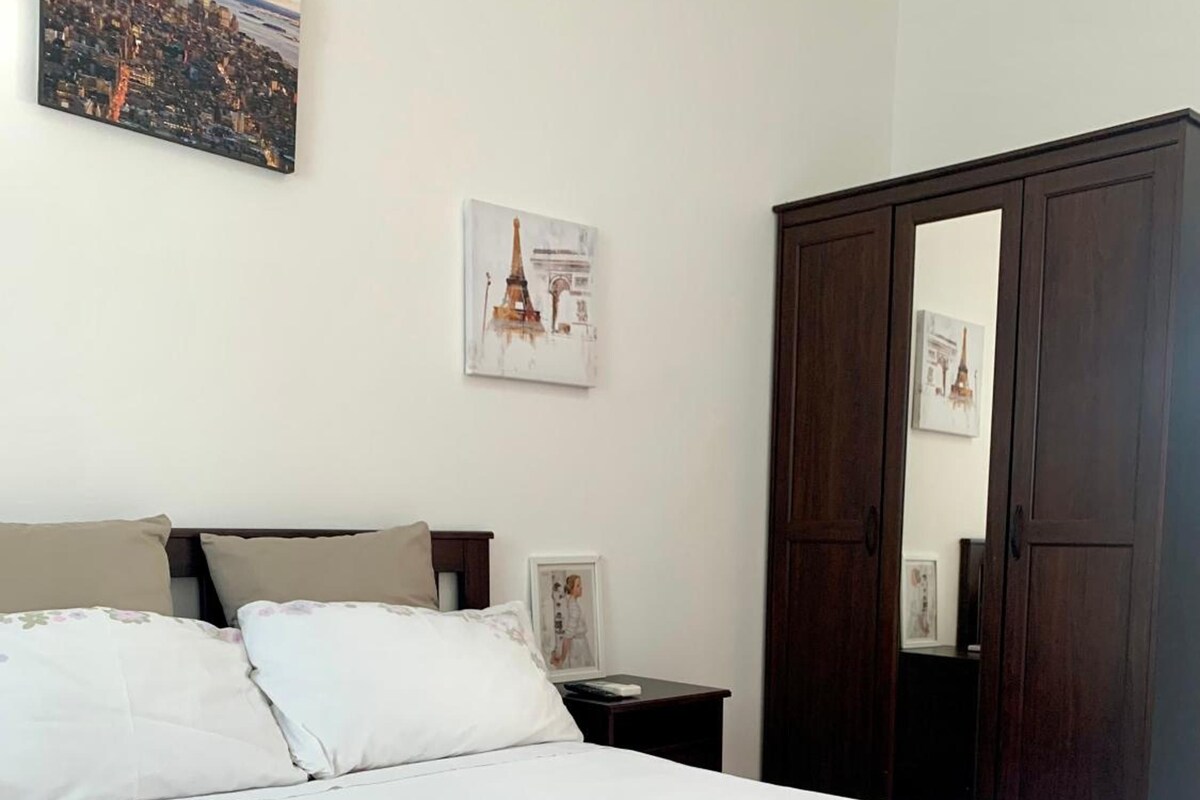 Un letto ben rifatto con copriletto bianco e cuscini, un comodino con un libro e una cornice, e un armadio capiente con uno specchio.