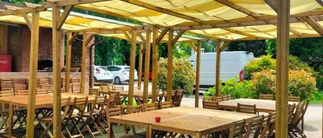 Un área de estar al aire libre con mesas y sillas de madera, cubierta por una pérgola con toldos amarillos.