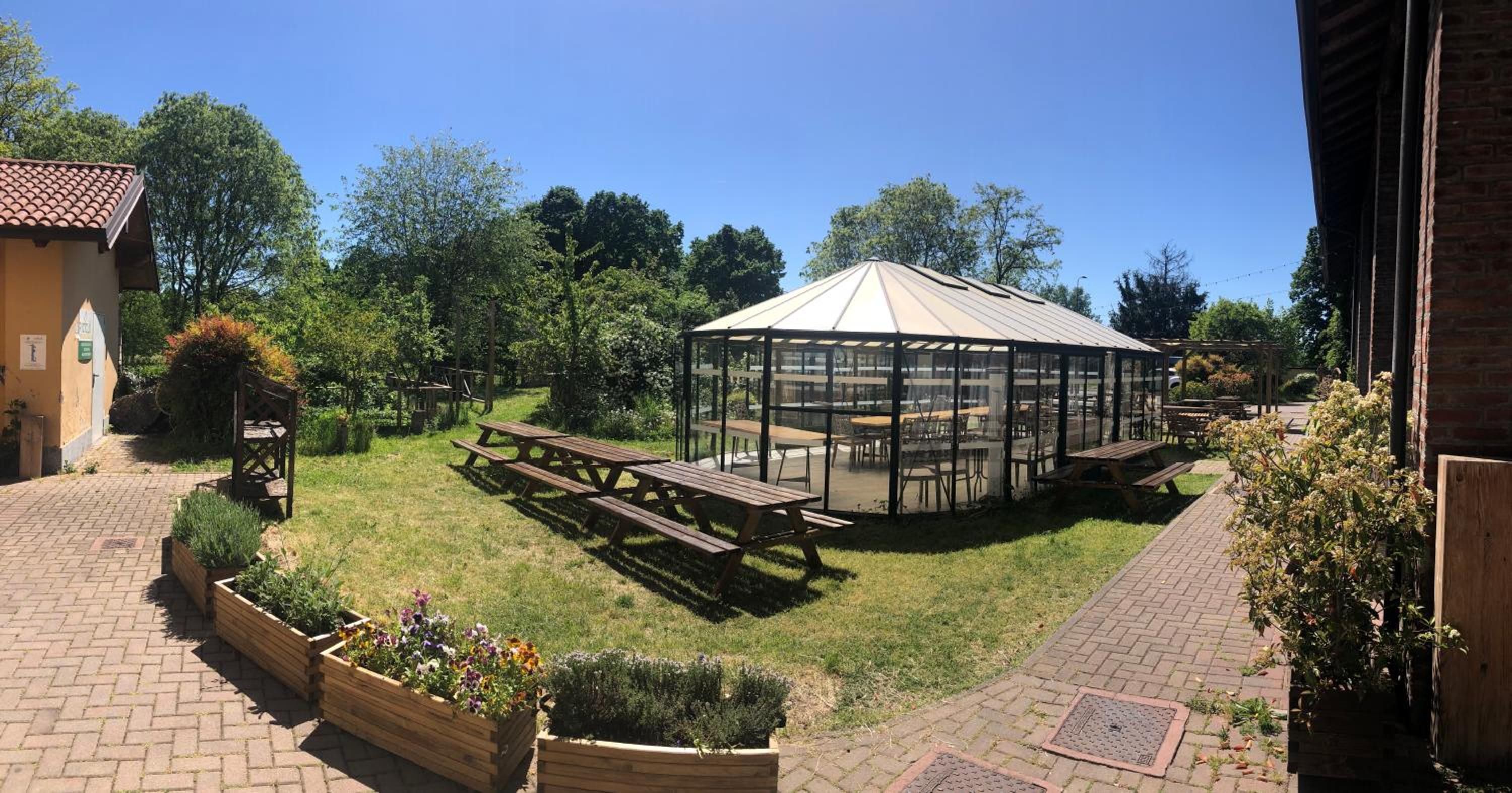 A gazebo with a transparent roof, surrounded by picnic tables and benches, set in a garden with brick pathways and wooden planters.