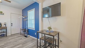 A room with a blue accent wall, a flat-screen TV mounted on the wall, a wooden table with a potted plant, and a door leading to another room. - Super Value Suite #6 - *4 km from FIFA Fan Fest* (Houston)