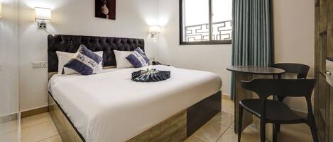 A hotel room with a large bed, a small table, two chairs, a window with curtains, and wall-mounted lights.