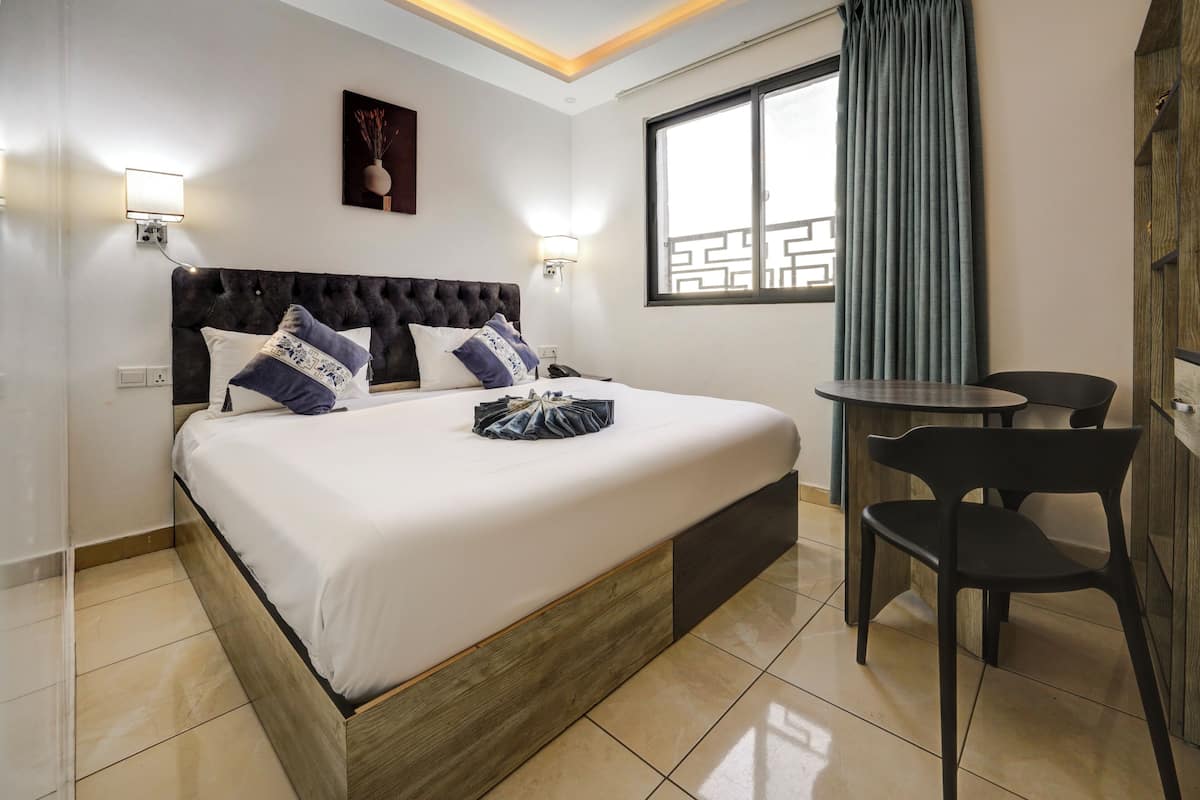 Une chambre d’hôtel avec un grand lit, une petite table, deux chaises, une fenêtre avec des rideaux et des lampes fixées au mur.