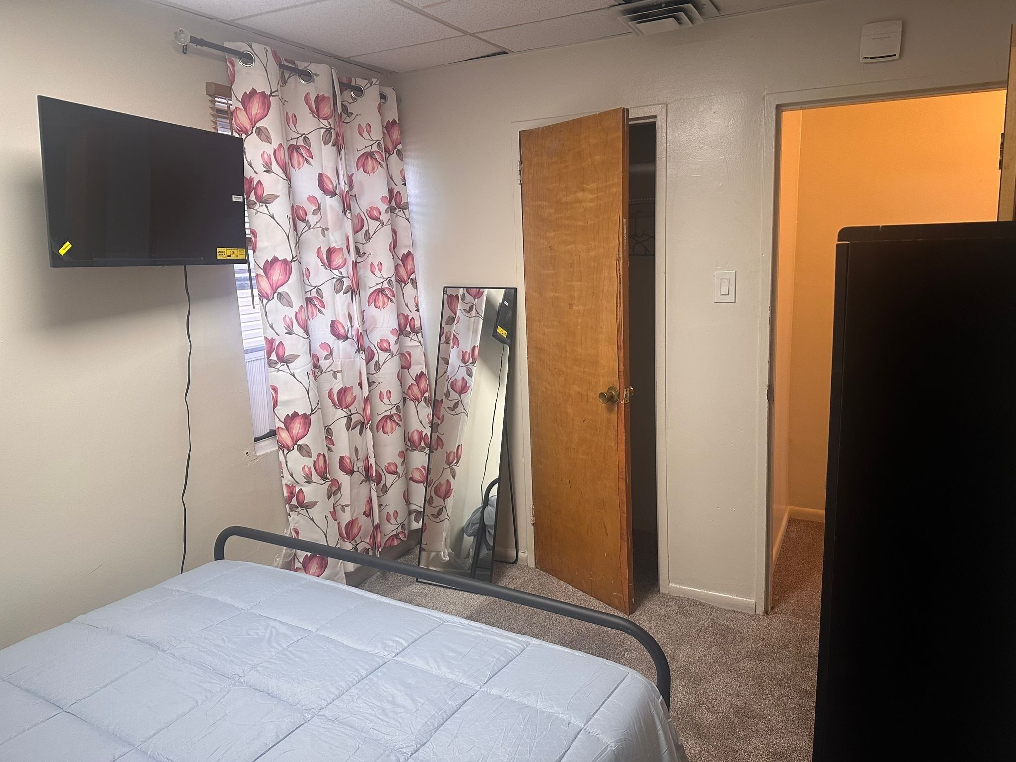 Une chambre à coucher avec un lit, un téléviseur fixé au mur, une porte donnant sur une autre pièce et une fenêtre ornée de rideaux à motifs floraux.