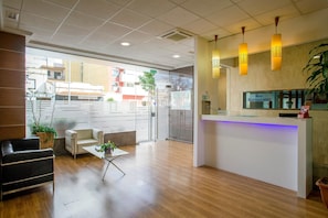 Un área de recepción con mostrador blanco, zona de estar con sofá y mesa de centro, y vista al exterior a través de un muro de cristal.