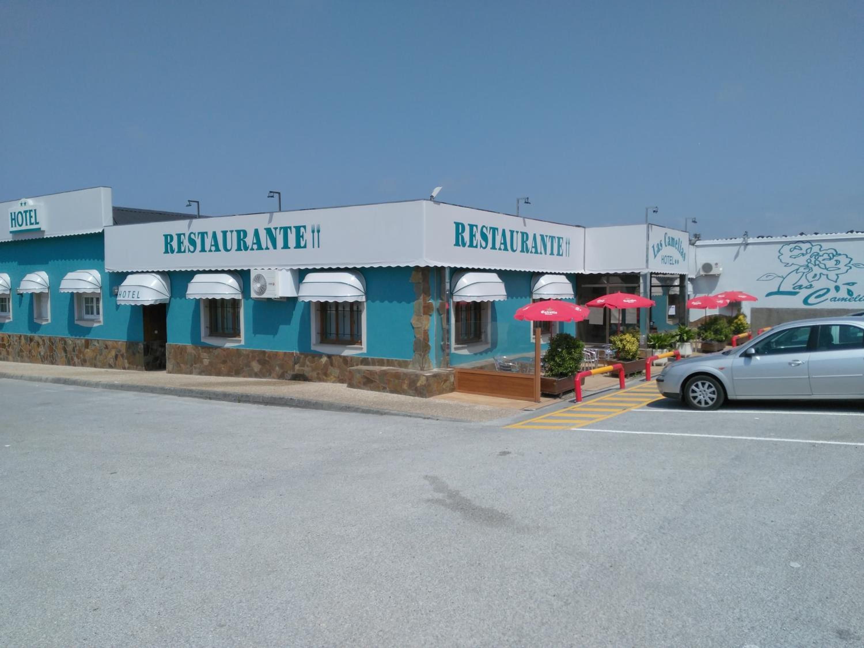 A building with a sign that reads 'RESTAURANTE' and 'HOTEL'. There are outdoor tables and chairs, and a car is parked in front.