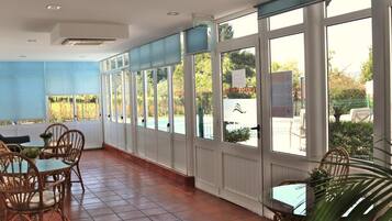 A covered outdoor seating area with tables and chairs, large windows, and a view of the outdoors.