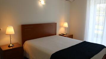 A hotel room with a single bed, two bedside tables with lamps, a wooden headboard, an air conditioning unit, and a window with sheer curtains.