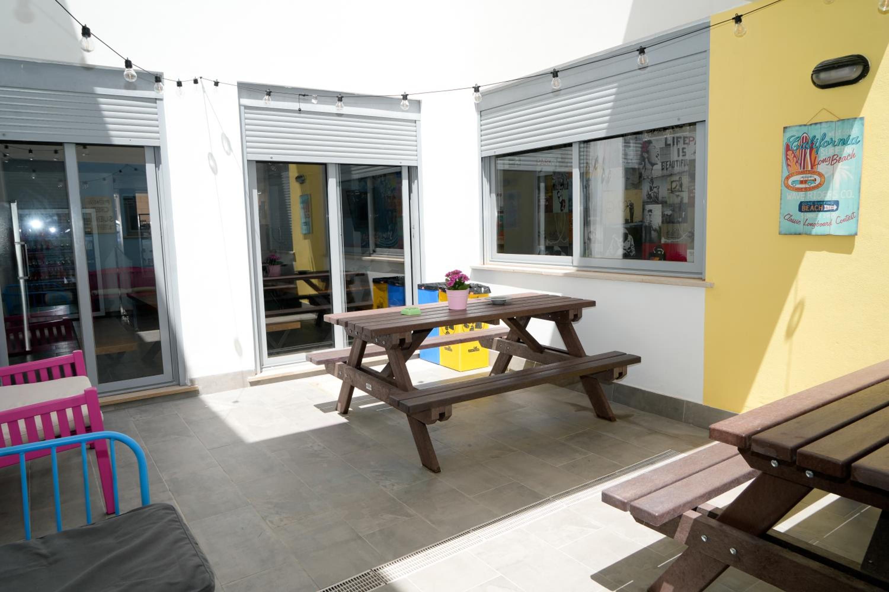 Un patio al aire libre con una mesa de picnic de madera, bancos y una pared amarilla con un cartel.