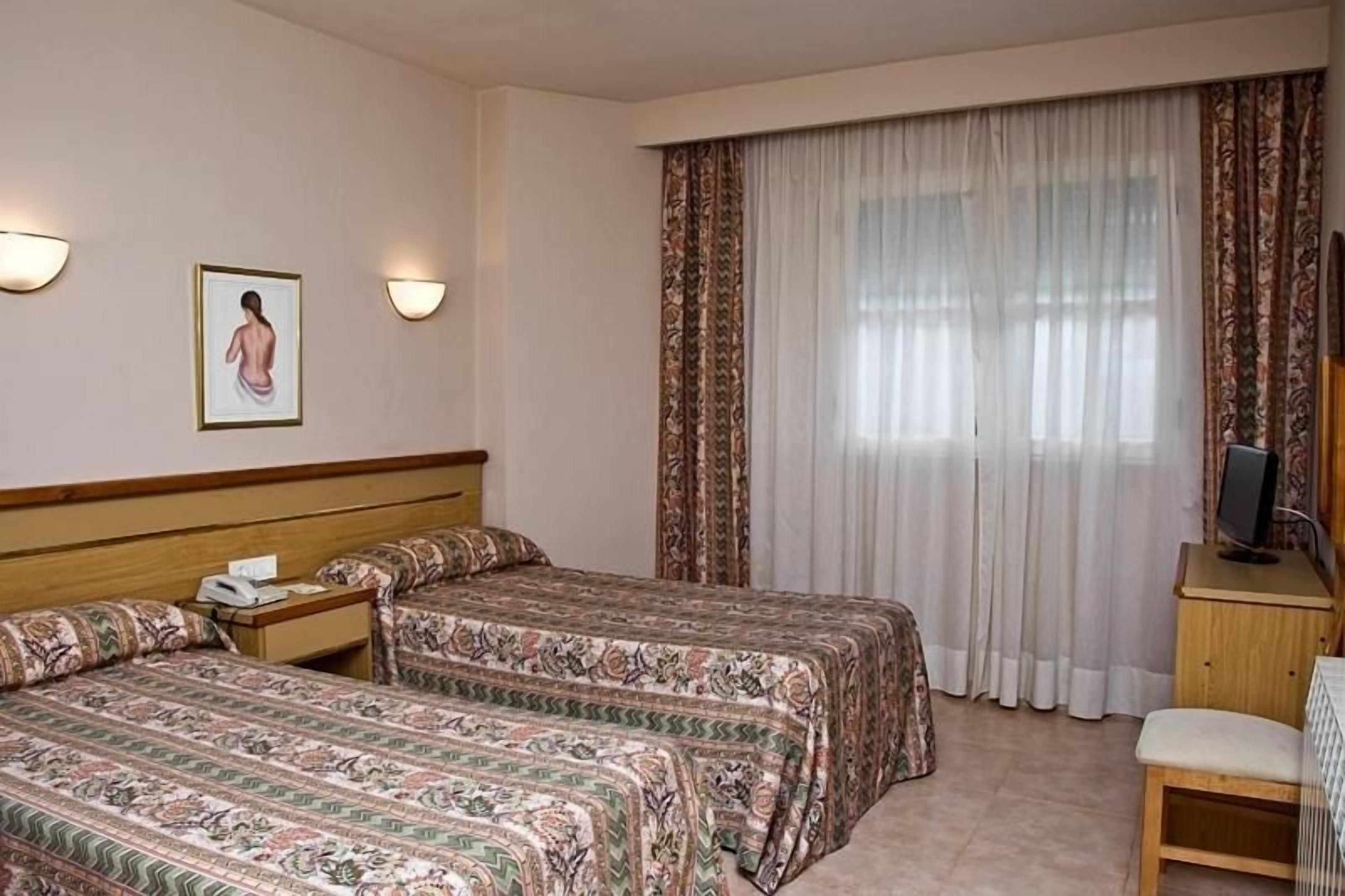 A hotel room with two beds, a desk, a chair, a television, and a window with curtains.
