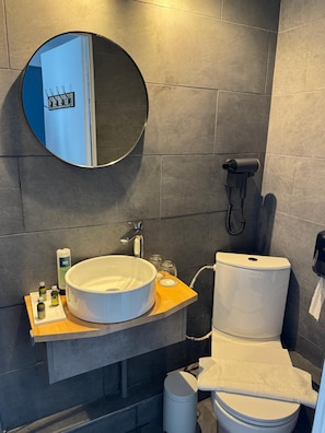 A modern bathroom with a round mirror, a white sink, a toilet, and a hairdryer on the wall. - Logis Hotel Spa La Poste (Vitry-le-Francois)