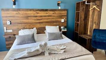 A hotel room with a wooden headboard, a bed with white linens and grey pillows, a blue chair, and a wooden wardrobe with a hanging rack.