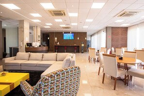 A hotel lobby with a sofa, chairs, a dining area, a reception desk, and a TV screen. - Tango Suites Cumbuco (Caucaia)
