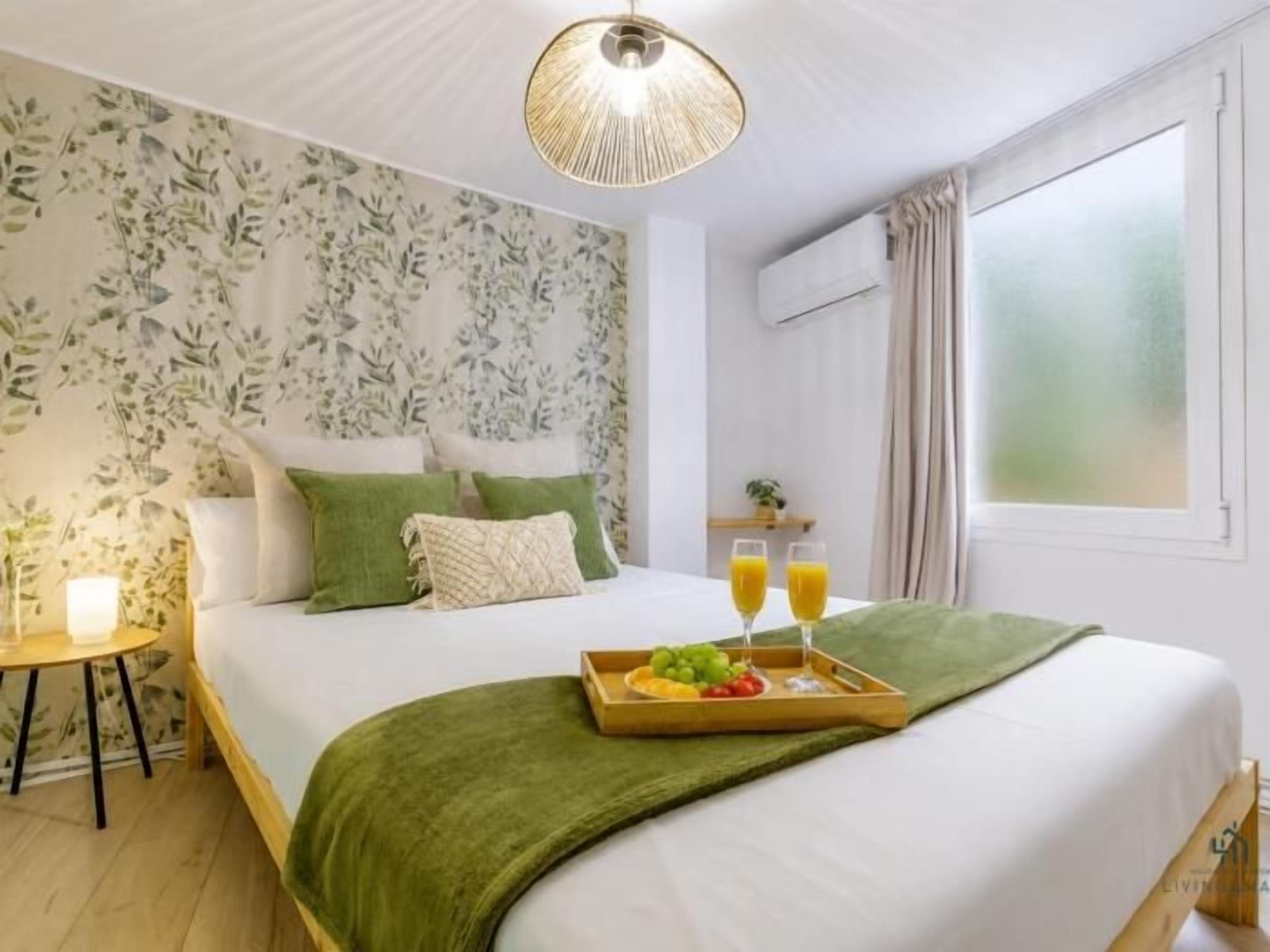 Une chambre d’hôtel avec un lit, une table de chevet avec une lampe, un plateau avec des fruits et du jus, et une fenêtre avec des rideaux.