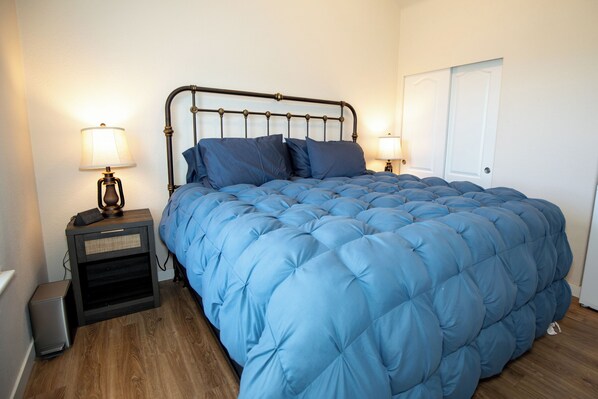 Un dormitorio con cama, dos mesitas de noche con lámparas, una puerta y un pequeño cubo de basura.