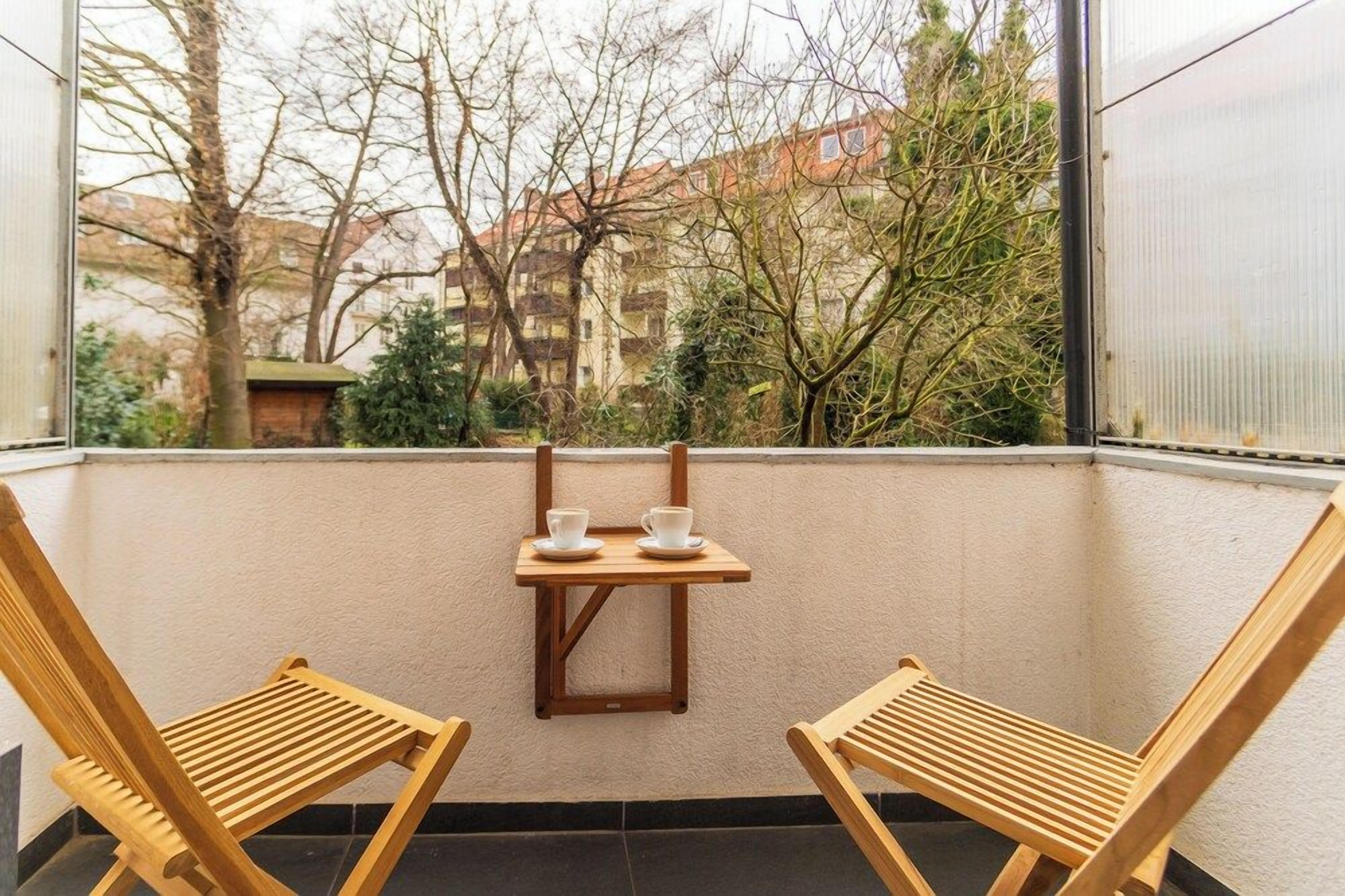A balcony with two wooden chairs and a small table with cups, overlooking a garden and buildings.