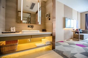 Ein modernes Badezimmer mit einem großen Spiegel, einem Waschbecken und einem Waschtisch. Unter dem Waschbecken befindet sich ein Korb und Handtücher liegen auf einem Regal.