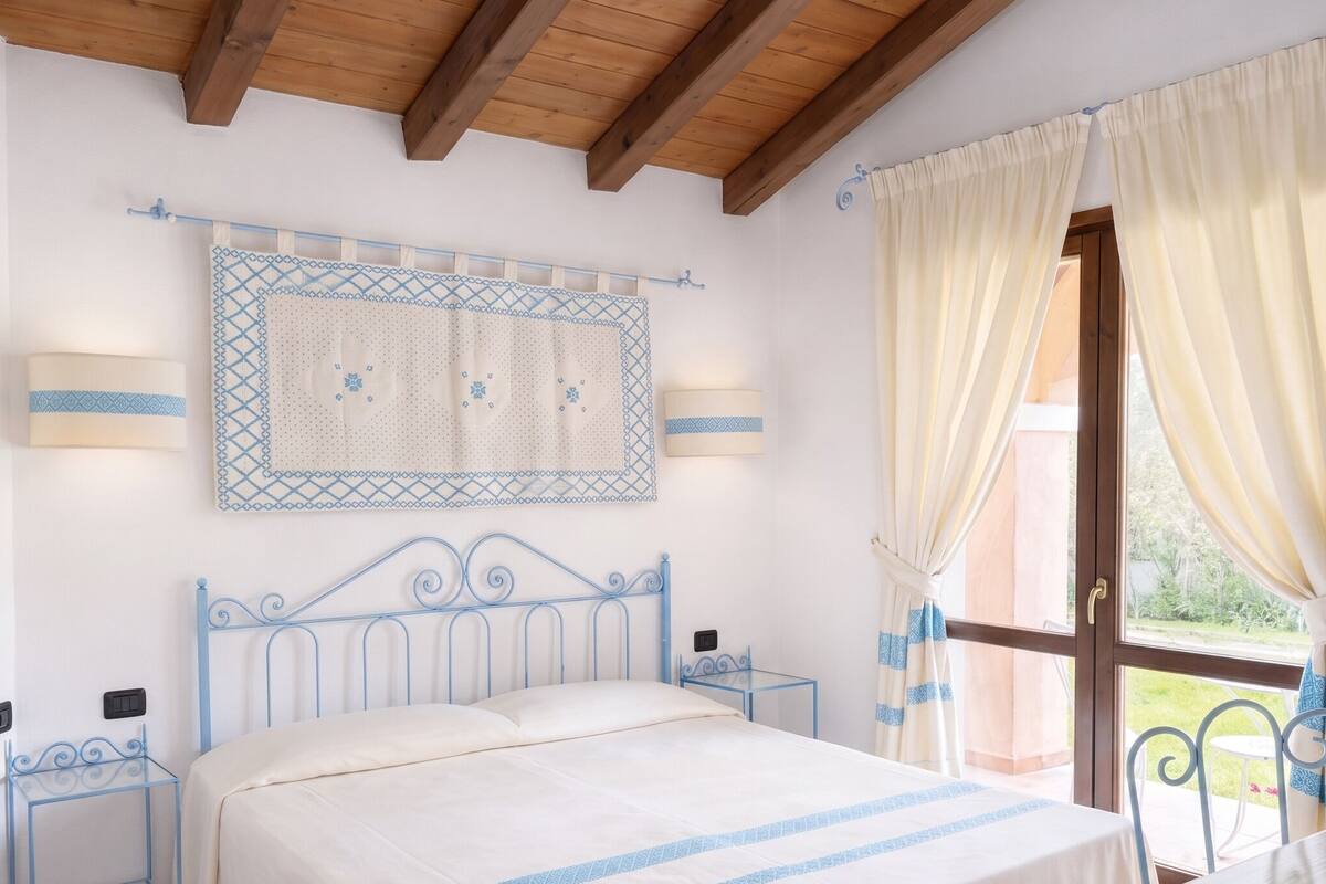 Un dormitorio con cama, mesitas de noche, una ventana con cortinas, un aplique de pared y un techo con vigas de madera.