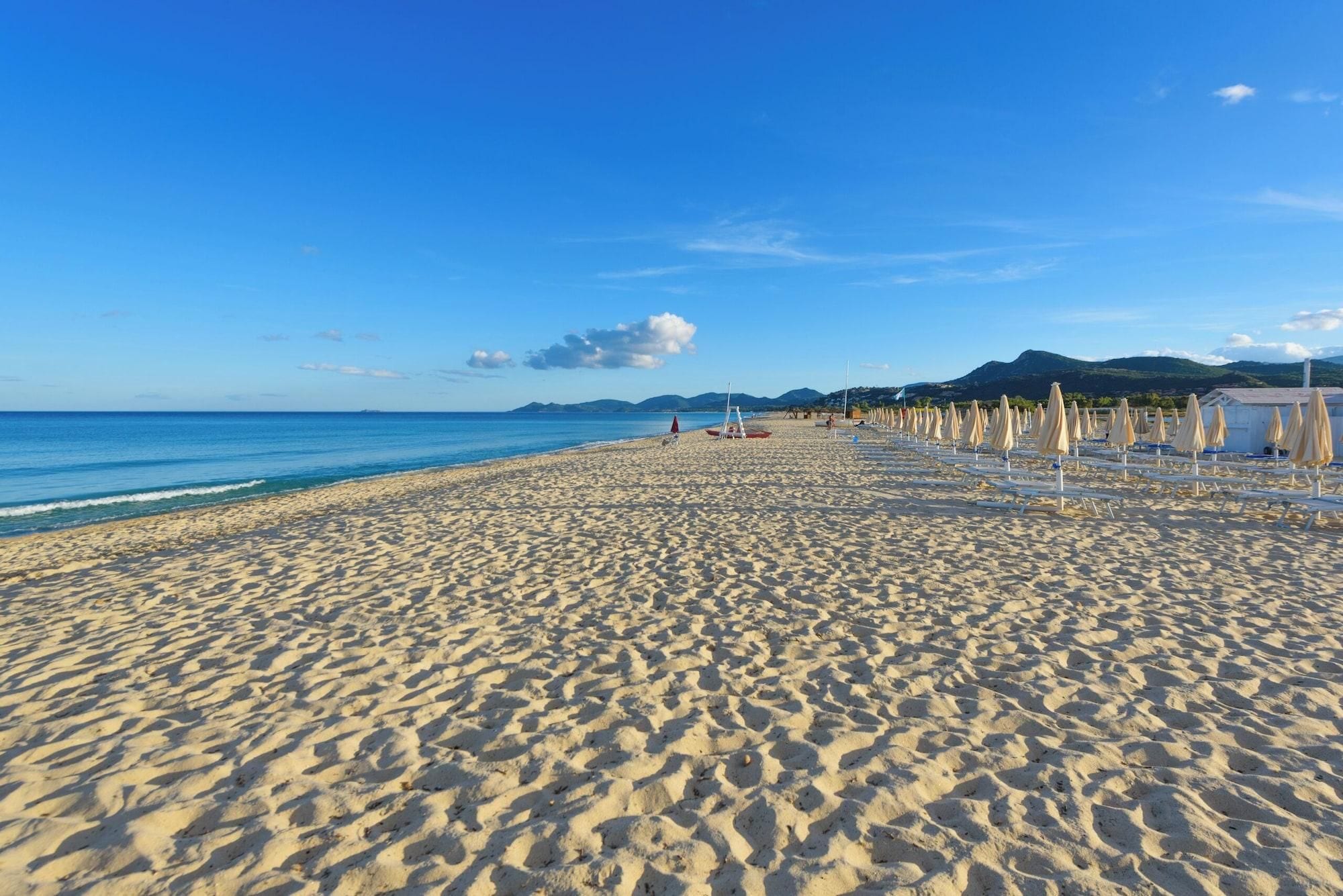 A wide sandy beach with rows of beach chairs and umbrellas, a calm sea, and distant mountains.