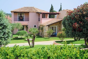 A two-story building with a stone facade, a balcony, and a well-maintained garden with various shrubs and a palm tree.