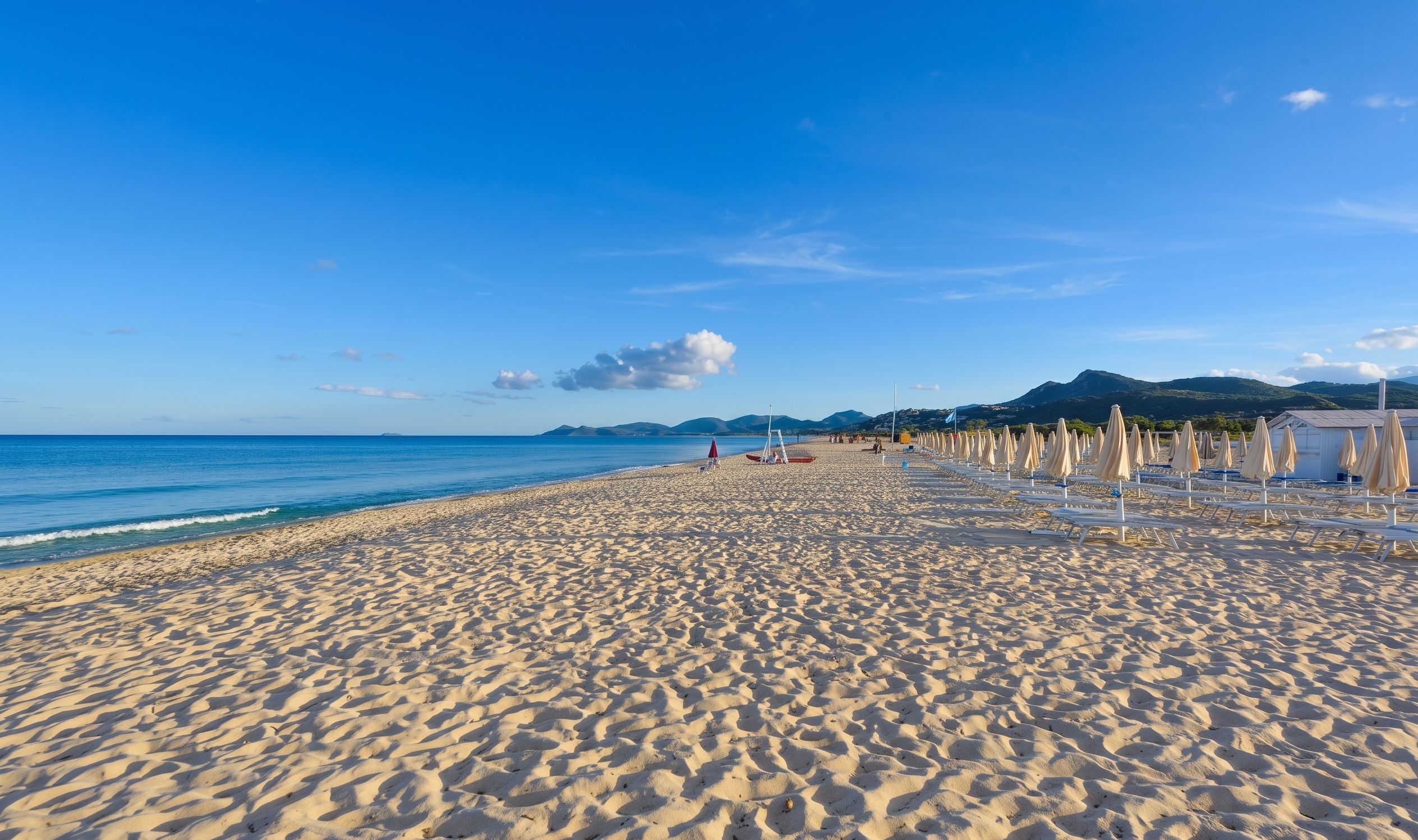 A wide sandy beach with rows of beach chairs and umbrellas, a clear blue sky, and distant mountains.