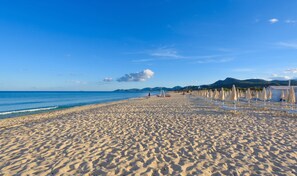 Ein weiter Sandstrand mit Liegestühlen und Sonnenschirmen, ein klarer blauer Himmel und ferne Berge.