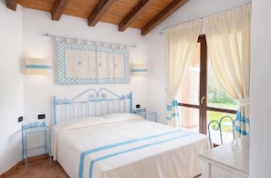 A bedroom with a bed, a nightstand, a wall-mounted lamp, a window with curtains, and a view of the outdoors.