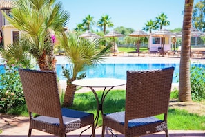 A poolside area with wicker chairs and a table, surrounded by palm trees and umbrellas.