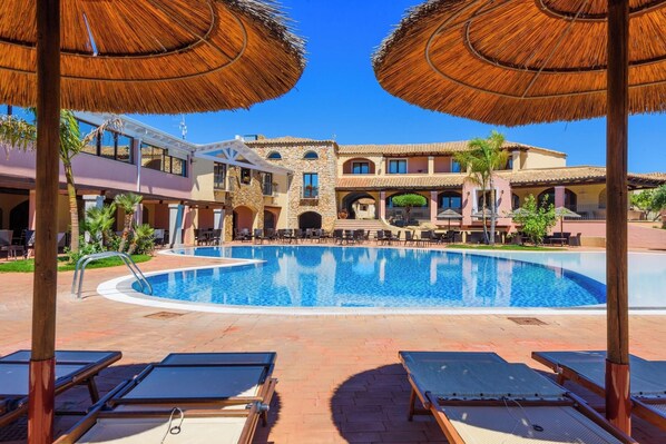 Un área de piscina con tumbonas y sombrillas, un edificio de piedra con balcones y un cielo azul despejado.