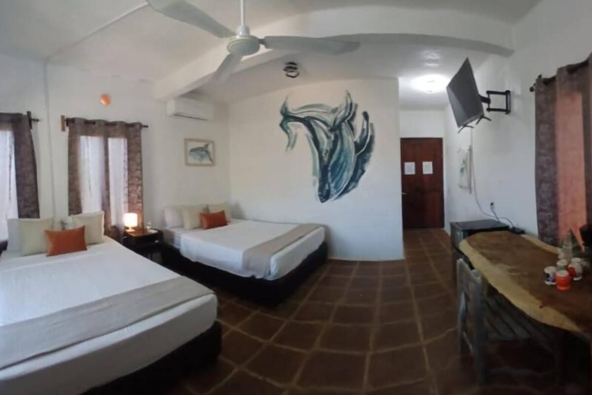 Une chambre d’hôtel avec deux lits, un ventilateur de plafond, une fresque murale, une table en bois et un téléviseur fixé au mur.