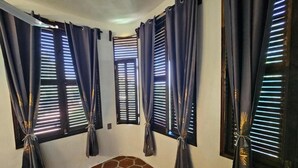 Una habitación con cuatro ventanas, cada una con cortinas oscuras atadas para mostrar persianas. El piso presenta un patrón hexagonal.