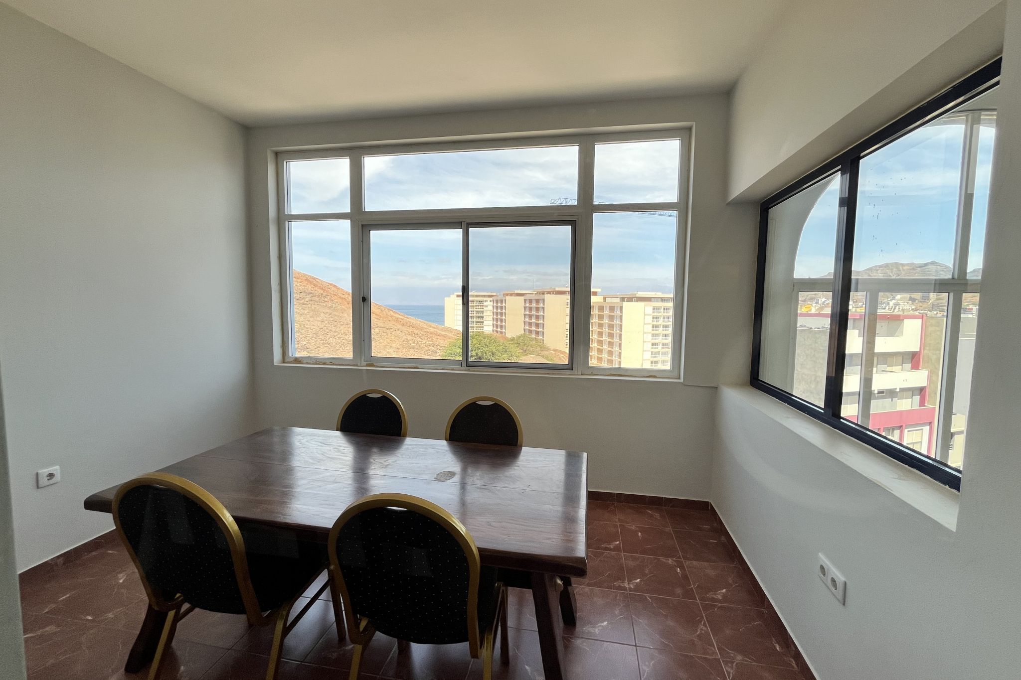 Un comedor con una mesa y sillas, ventanas amplias con vistas a edificios y colinas.