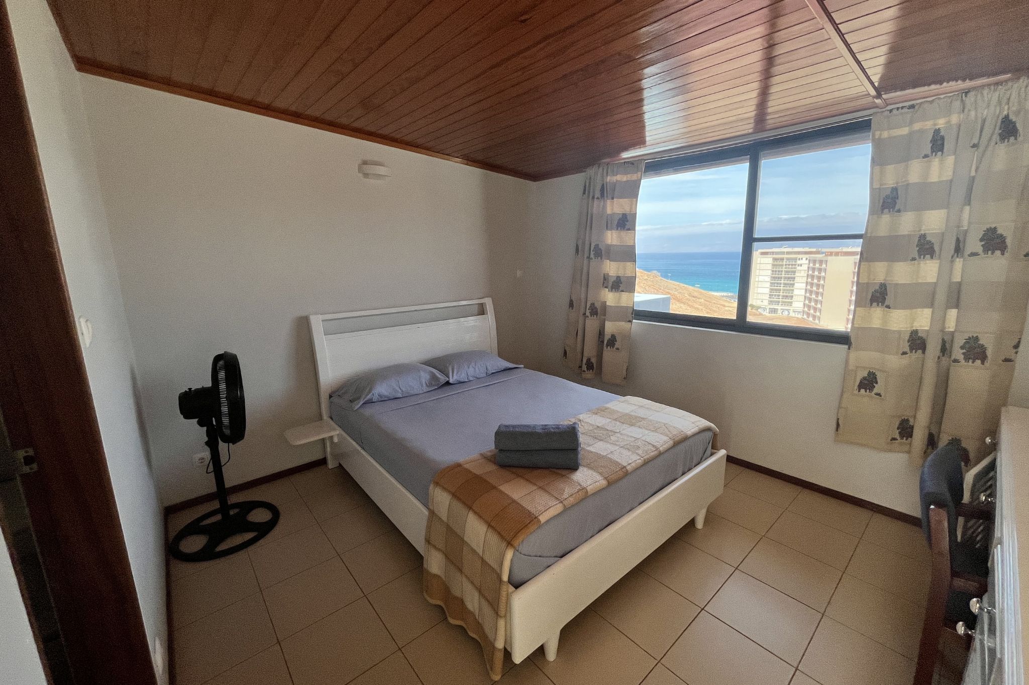 A bedroom with a bed, a standing fan, a window with a view, and a curtain with a pattern.