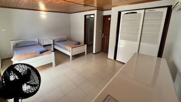 Habitación pequeña con dos camas individuales, una mesita, un ventilador y un armario con puertas persianas.